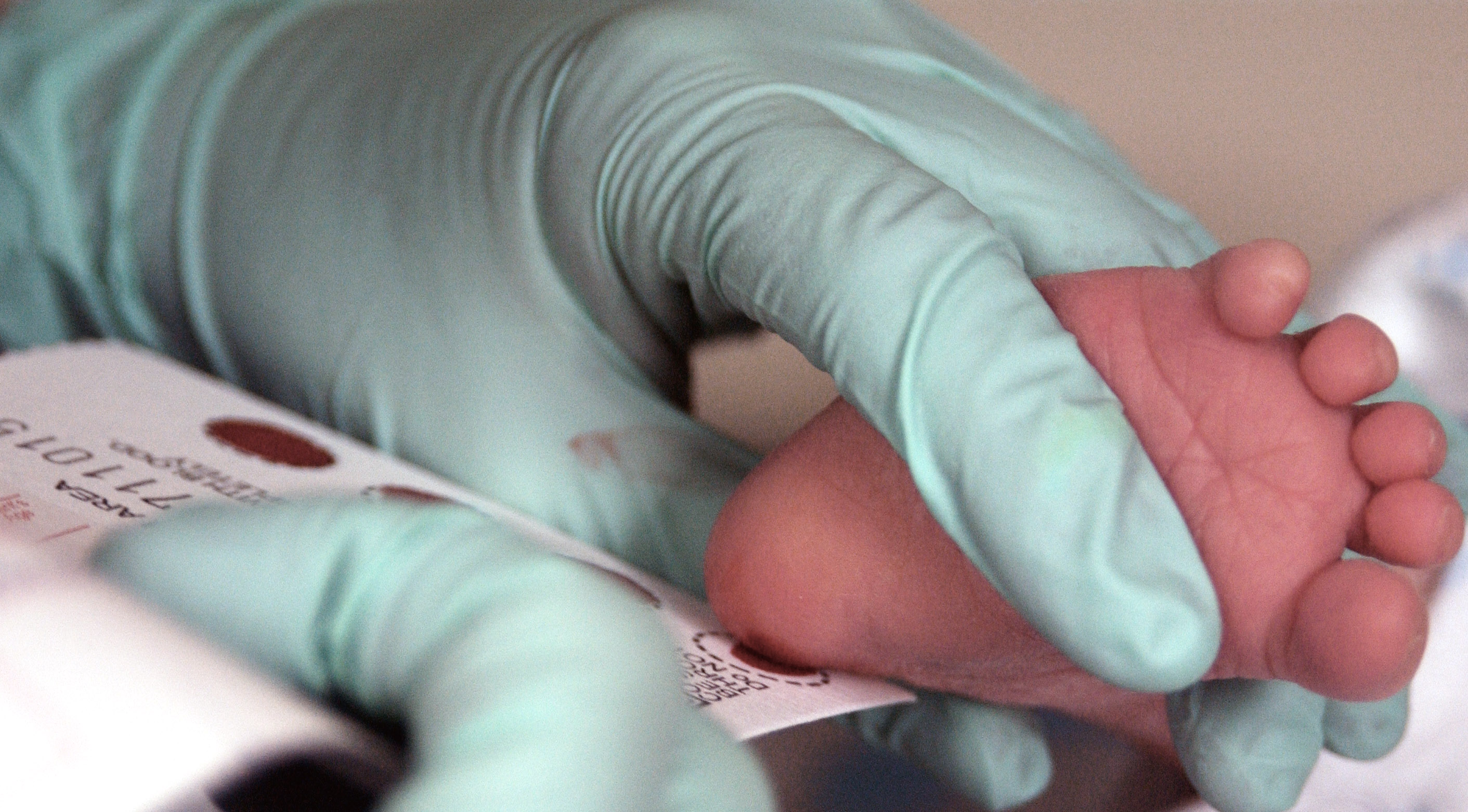 Hand in light blue glove holds newborn’s heel to a white card, which absorbs a spot of blood.