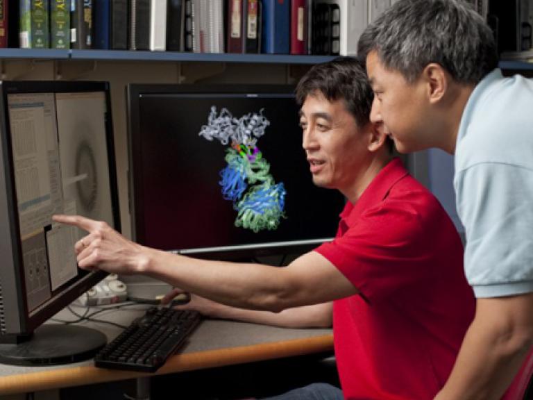 Profile picture of Peter Kwong and Tongqing Zhou working at a computer. Peter Kwong is seated and pointing at the monitor screen, while Tongqing looks on behind him.