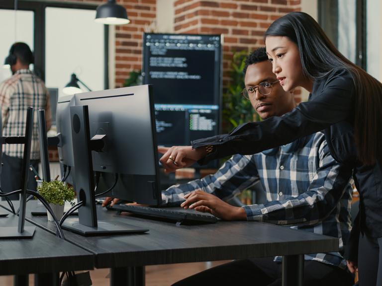 two people working together at a computer, a person with virtual reality goggles on in the background