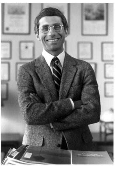 A black and white photo of Dr. Fauci standing with arms crossed in front of a wall with various plaques and frames