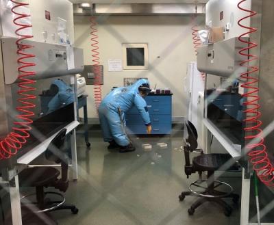 In a laboratory environment, a woman wearing a blue personal protective equipment suit bends over to pick up two small white boxes.