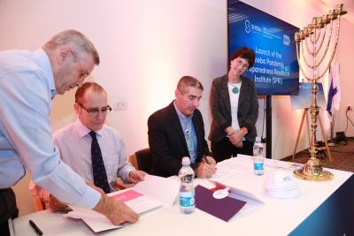 A man and a woman stand on either side of a table as a seated man signs a document. The man seated on his right looks on.  