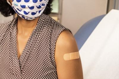 Woman wearing a face mask sitting in a doctors office with a bandage placed over an injection site on her arm.