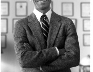 A black and white photo of Dr. Fauci standing with arms crossed in front of a wall with various plaques and frames