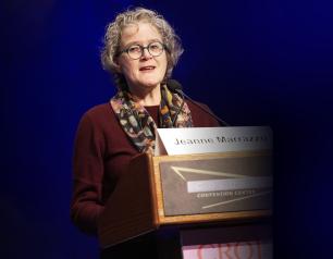 A person stands at a podium with "Jeanne Marrazzo" name card and "CROI" banner below.